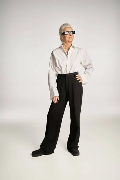 Жінка середнього віку в сонцезахисних окулярах і елегантний одяг, що стоїть з рукою на стегні на сірому тлі — стокове фото