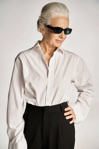 Middle aged woman in sunglasses and elegant attire standing with hand on hip on grey backdrop — Stock Photo