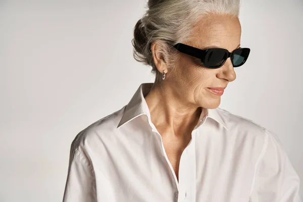 Portrait of stylish middle aged woman in white shirt and sunglasses posing on grey background — Stock Photo