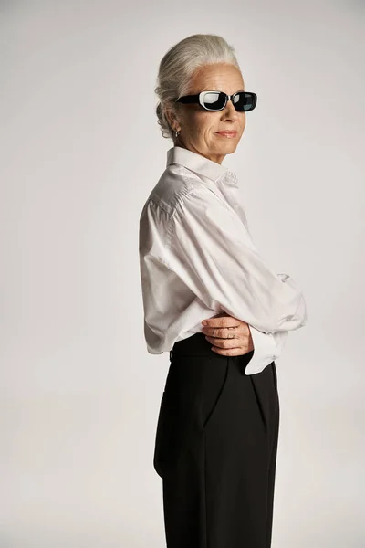Porträt einer eleganten Frau mittleren Alters in weißem Hemd und Sonnenbrille, die auf grauem Hintergrund posiert — Stockfoto