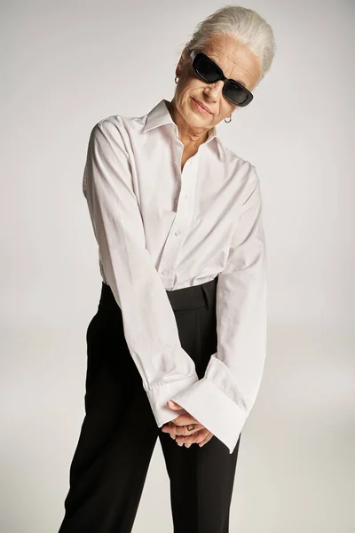 Fashionable middle aged woman in white shirt, black pants and sunglasses posing on grey backdrop — Stock Photo