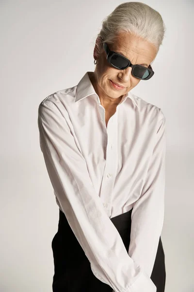 Fashionable middle aged woman in white shirt, black pants and sunglasses posing on grey backdrop — Stock Photo