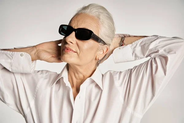 Portrait of fashionable middle aged woman in white shirt and sunglasses posing on grey backdrop — Stock Photo