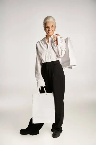 Stylish middle aged woman in classy white shirt and black pants standing with shopping bags on grey — Stock Photo