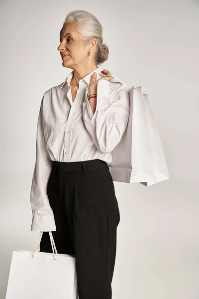Elegante donna di mezza età in camicia bianca di classe e pantaloni neri in piedi con borse della spesa su grigio — Foto stock
