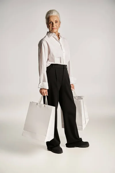 Full length, middle aged woman in white shirt and black pants standing with shopping bags on grey — Stock Photo