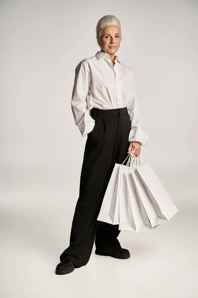Donna di mezza età in camicia bianca e pantaloni in piedi con mano in tasca e tenendo borse della spesa — Foto stock