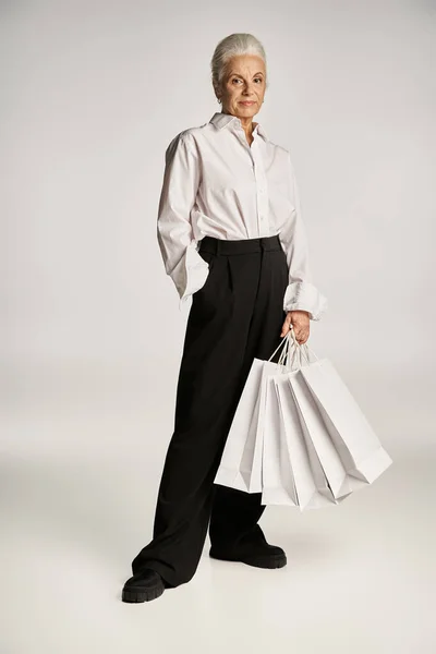 Middle aged woman in white shirt and pants standing with hand in pocket and holding shopping bags — Stock Photo