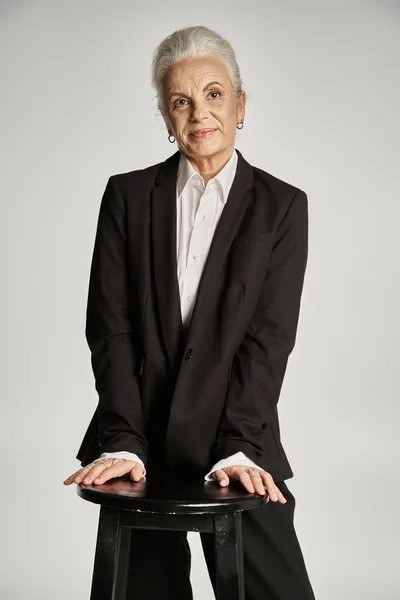 Hermosa y gris mujer de mediana edad en traje elegante posando sobre fondo gris - foto de stock