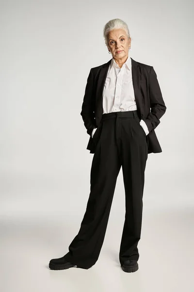 Hermosa y gris mujer de mediana edad en traje elegante posando sobre fondo gris - foto de stock