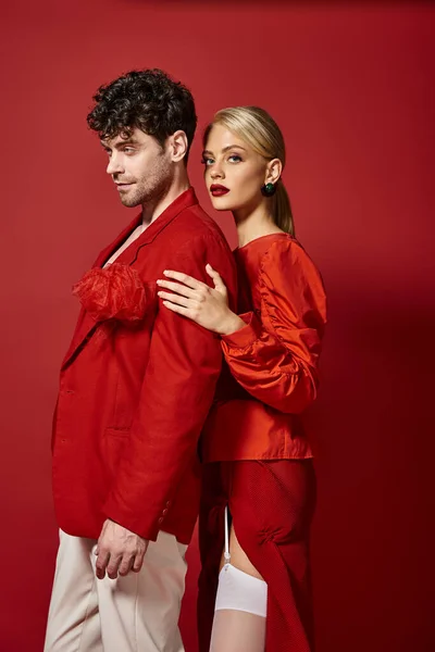 Mulher loira com lábios vermelhos abraçando homem bonito em trajes vibrantes de fundo vermelho, moda — Fotografia de Stock