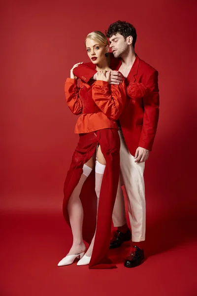 Handsome man embracing blonde woman in elegant attire on red background, fashionable couple — Stock Photo