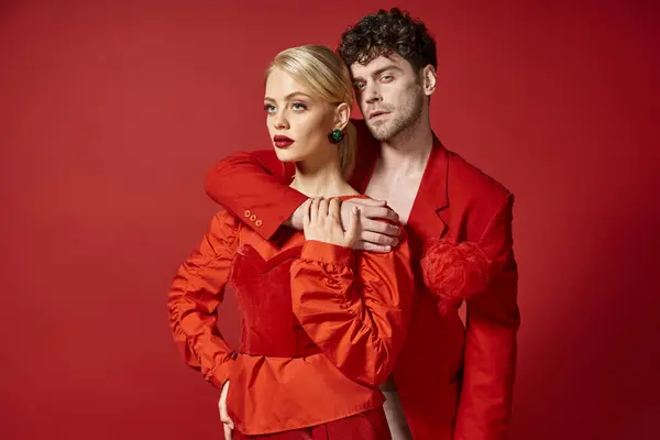 Schöner Mann umarmt schöne Frau in eleganter Kleidung auf rotem Hintergrund, modisches Paar — Stockfoto