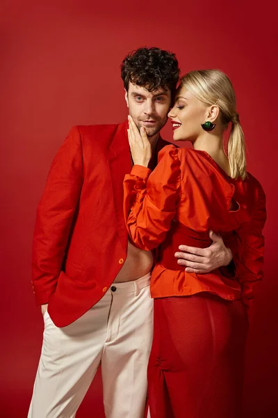 Mujer rubia feliz con labios rojos tocando la cara del hombre guapo en traje elegante en el telón de fondo vibrante - foto de stock