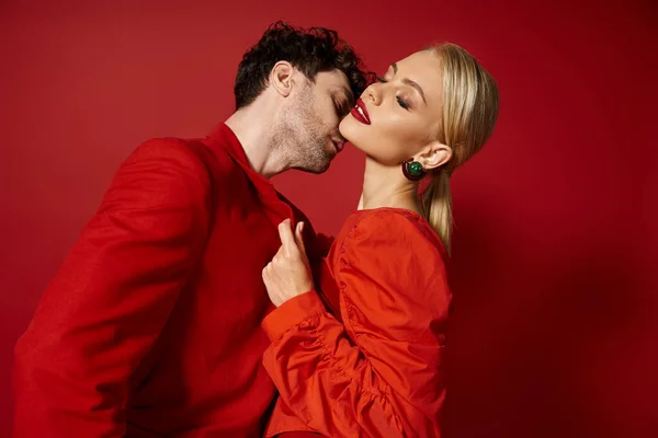 Mujer rubia en vestido y pendientes rojos seduciendo hombre guapo sobre fondo rojo, pareja de moda - foto de stock