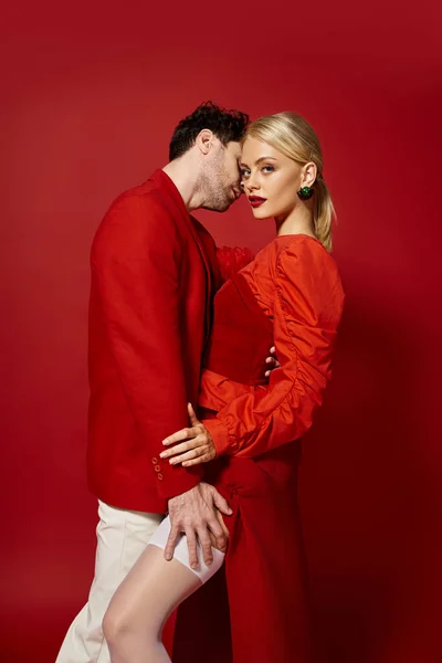 Hombre guapo tocando pierna de mujer rubia seductora en medias sexy sobre fondo rojo, moda - foto de stock