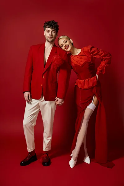 Glückliche blonde Frau hält die Hände eines gutaussehenden Mannes in stilvoller Kleidung auf rotem Hintergrund, Mode — Stockfoto
