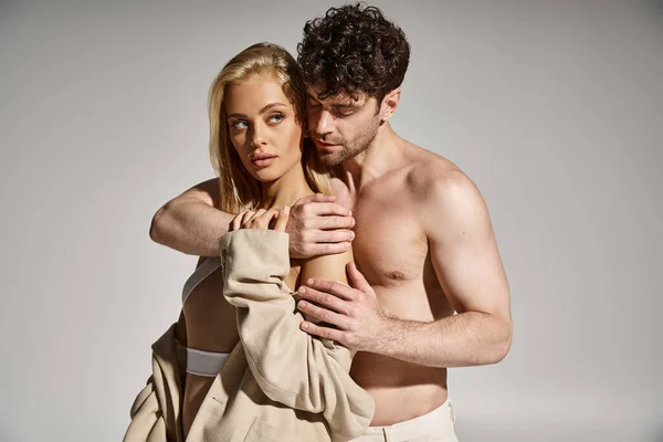 Mann ohne Hemd posiert mit schöner, blonder Frau im Body und beigem Blazer auf grauem Hintergrund — Stockfoto
