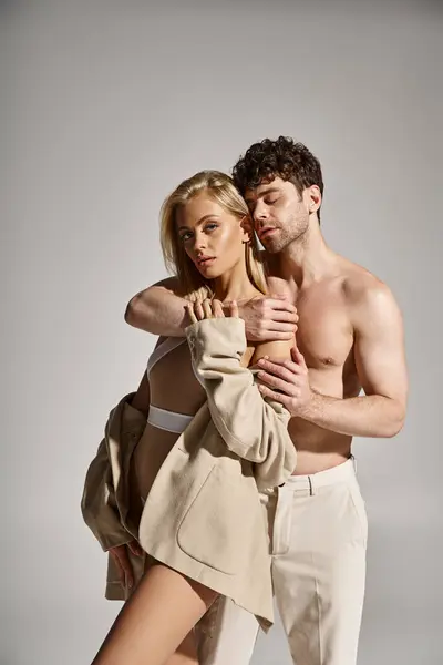 Hombre sin camisa posando con mujer encantadora y rubia en body y blazer beige sobre fondo gris - foto de stock
