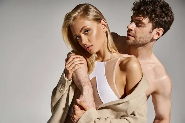 Mann ohne Hemd posiert mit hübscher Frau im Body und stilvollem beigen Blazer auf grauem Hintergrund — Stockfoto