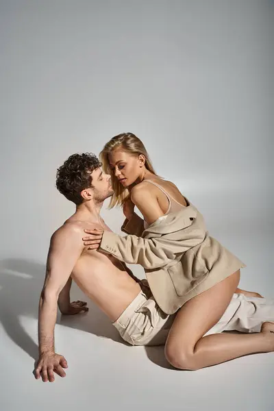Сексуальная блондинка в бежевом блейзере соблазняет красивого мужчину без рубашки на сером фоне, горячая пара — стоковое фото