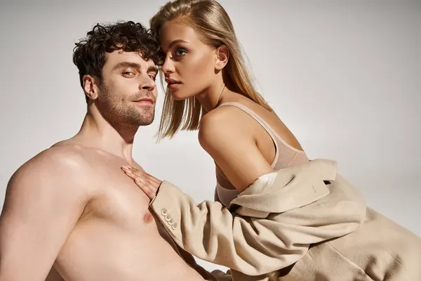 Junge blonde Frau im beigen Blazer verführt gutaussehenden hemdlosen Mann auf grauem Hintergrund, sexy Paar — Stockfoto