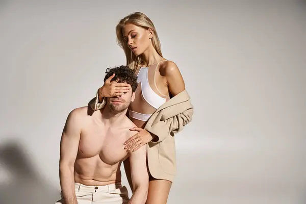 Mujer rubia en traje sexy cubriendo los ojos de hombre guapo sin camisa sobre fondo gris, pareja caliente - foto de stock