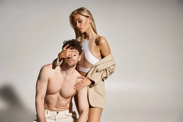 Blonde woman in sexy attire covering eyes of shirtless handsome man on grey background, hot couple — Stock Photo