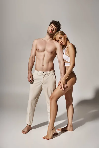 Hombre guapo con cuerpo musculoso cogido de la mano de mujer rubia bonita sobre fondo gris, sexy pareja - foto de stock