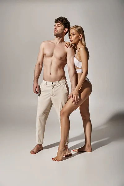 Без сорочки чоловік з м'язистим тілом тримає руку молодої блондинки на сірому фоні, сексуальна пара — стокове фото
