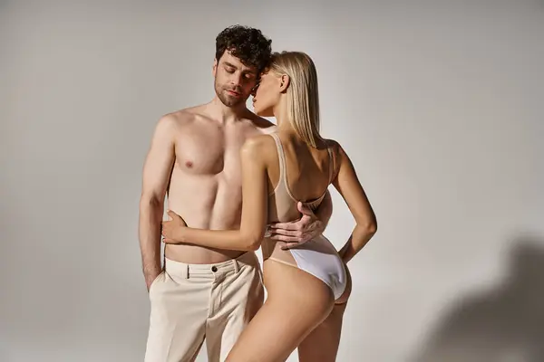 Schöner Mann mit muskulösem Körper verführt blonde Frau im Body auf grauem Hintergrund, sexy Paar — Stockfoto