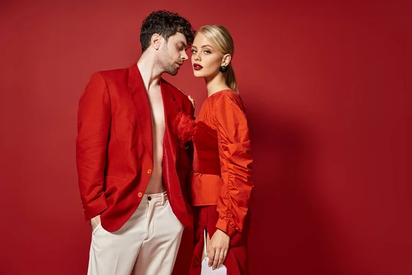 Hombre guapo en traje de diseñador rojo posando con modelo rubio sobre un fondo vibrante, pareja de moda - foto de stock