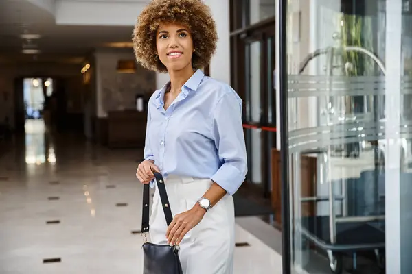Elegantemente vestida y feliz mujer afroamericana con bolso de pie en el vestíbulo del hotel - foto de stock
