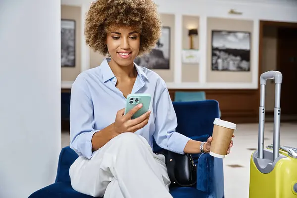 Relajada y rizada mujer afroamericana con café para llevar y teléfono inteligente esperando en el vestíbulo del hotel - foto de stock
