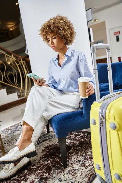 Relajada y feliz mujer afroamericana con café para llevar y teléfono inteligente esperando en el vestíbulo del hotel - foto de stock