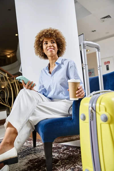 Femme afro-américaine détendue et heureuse avec café à emporter et smartphone en attente dans le hall de l'hôtel — Photo de stock