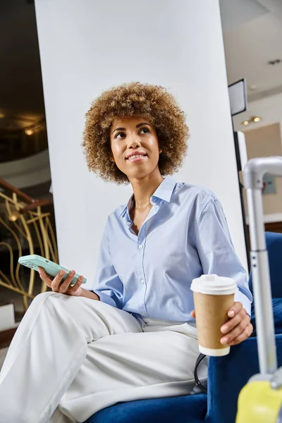 Mujer afroamericana relajada y soñadora con café para llevar y teléfono inteligente esperando en el vestíbulo del hotel - foto de stock
