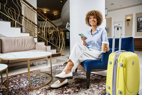 Relaxed and happy african american woman with coffee to go and smartphone sitting in hotel lobby — Stock Photo
