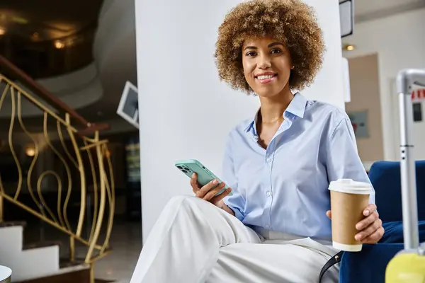 Mujer afroamericana rizada y feliz con café para llevar y teléfono inteligente esperando en el vestíbulo del hotel - foto de stock