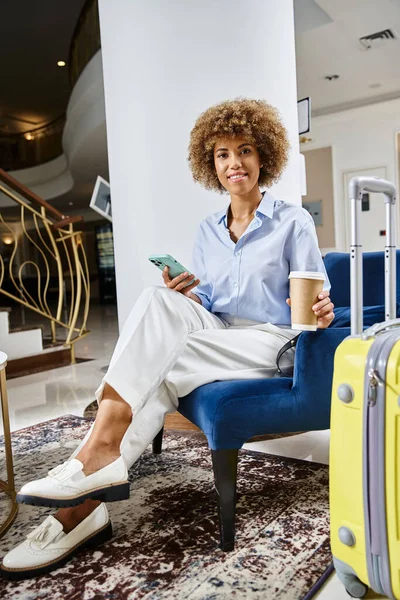 Mujer afroamericana relajada y sonriente con café para llevar y teléfono inteligente esperando en el vestíbulo del hotel - foto de stock