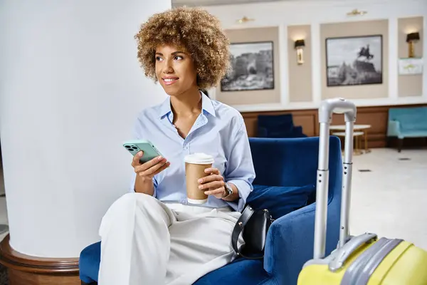Relajada y sonriente mujer afroamericana con café para llevar y teléfono inteligente sentado en el vestíbulo del hotel - foto de stock