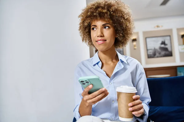 Happy curly african american woman with coffee to go and smartphone sitting in hotel lobby — Stock Photo