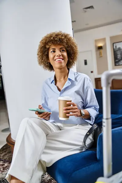 Alegre rizado mujer afroamericana con café para llevar y el uso de teléfono inteligente en el vestíbulo del hotel - foto de stock