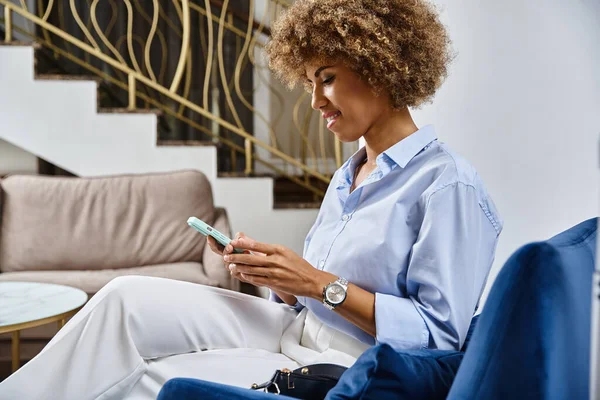 Mujer afroamericana de pelo rizado sonriendo y mensajes de texto en el teléfono inteligente en el vestíbulo del hotel, viajes - foto de stock