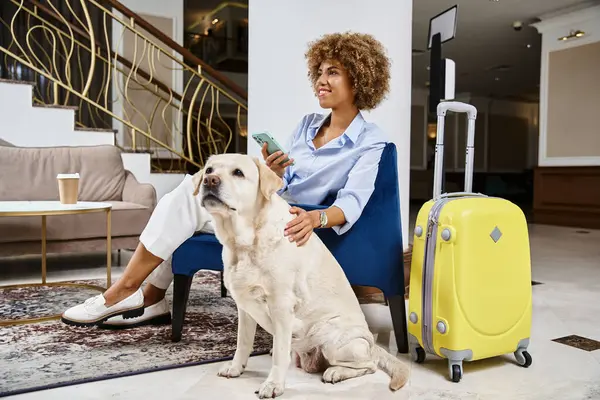 Mujer afroamericana sonriente con teléfono inteligente sentado cerca de labrador en el vestíbulo de un hotel que acepta mascotas - foto de stock