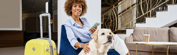 Mujer afroamericana feliz con teléfono inteligente sentado cerca de labrador en hotel que acepta mascotas, pancarta - foto de stock