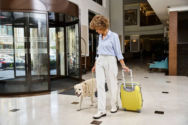 Turista feliz con su Labrador en una entrada de hotel que acepta mascotas, mujer afroamericana con perro - foto de stock