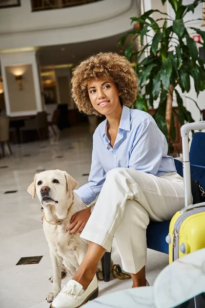 Feliz huésped esperando el check-in con Labrador en hotel que acepta mascotas, mujer negra abrazando perro - foto de stock