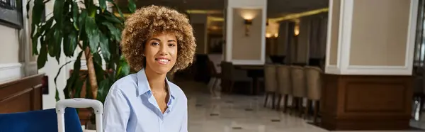 Mujer afroamericana feliz con el pelo rizado esperando el cheque en el vestíbulo del hotel, pancarta horizontal - foto de stock