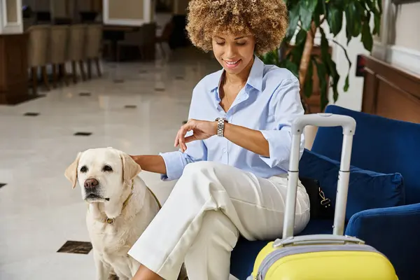 Feliz huésped a la espera de check-in con perro en hotel que acepta mascotas, mujer negra mirando reloj de pulsera - foto de stock